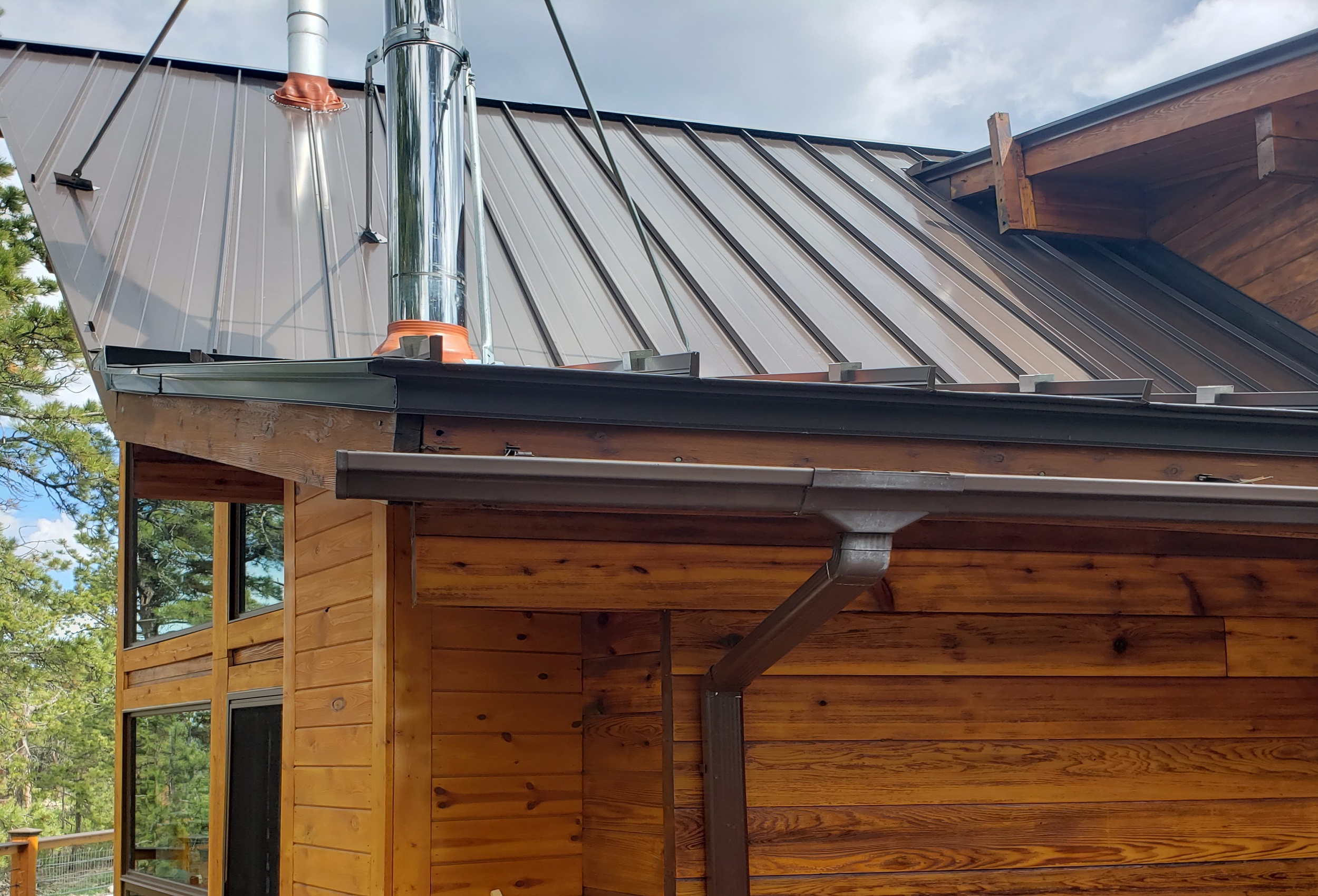 Seamless gutter installation on a metal roof cabin in Fort Collins, CO, with a downspout mounted along cedar siding